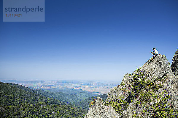 Männlicher Wanderer sitzt auf Fels vor klarem blauen Himmel