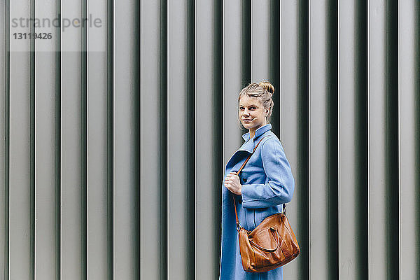 Seitenansicht Porträt einer selbstbewussten jungen Frau in Trenchcoat  die in der Stadt ihre Geldbörse gegen den Fensterladen drückt