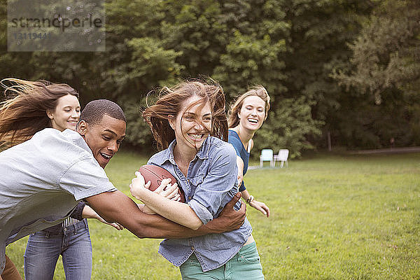 Mann zieht Frau mit dem Fussball in der Hand  während Freunde im Hintergrund auf dem Feld rennen
