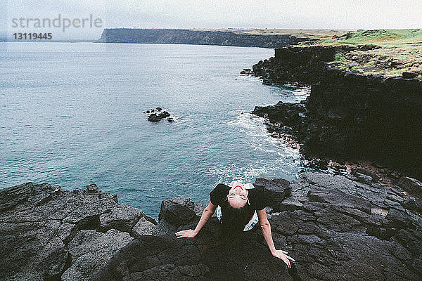 Hochwinkelansicht einer glücklichen Frau  die sich auf Felsen am Strand gegen den Himmel entspannt