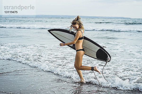 Seitenansicht einer Frau  die beim Strandspaziergang am Ufer ein Surfbrett trägt