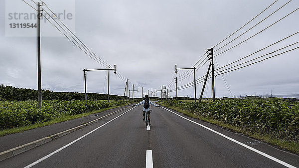 Rückansicht eines Mannes mit ausgestreckten Armen  der einen Rucksack trägt  während er mit dem Fahrrad auf der Straße vor bewölktem Himmel fährt