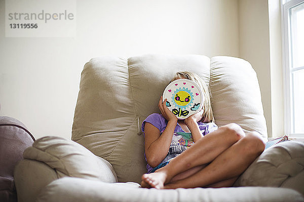 Mädchen hält Spielzeug an ihr Gesicht  während sie auf der Couch an der Wand sitzt