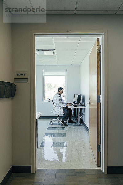 Seitenansicht eines Arztes  der einen Desktop-Computer benutzt  während er im Krankenhaus arbeitet  durch die Tür gesehen
