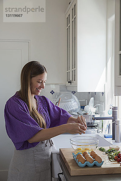Glückliche Frau zerbricht Eier in Rührschüssel  während sie in der Küche steht