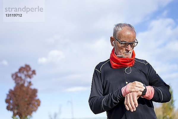 Ernsthafter älterer Mann mit Kopfhörern  der auf die Armbanduhr schaut  während er gegen den bewölkten Himmel im Park trainiert