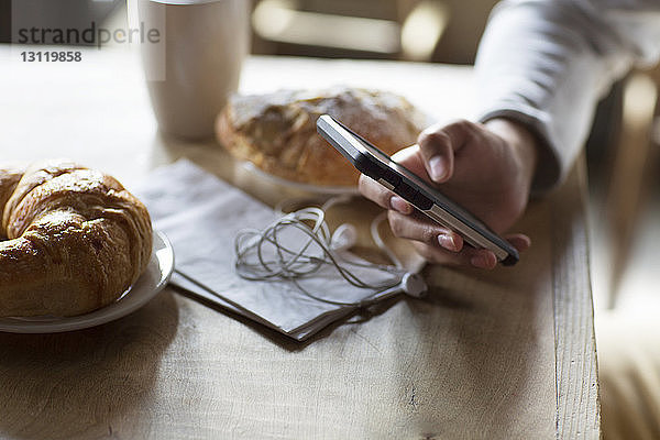 Ausgeschnittenes Bild eines Mannes  der sein Mobiltelefon bei Croissants und Kaffee am Tisch hält