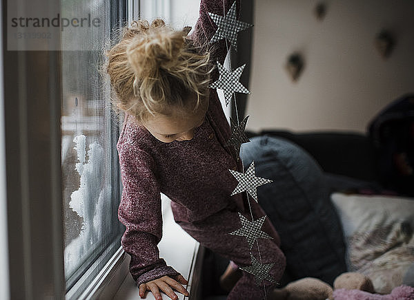 Mädchen hält sternförmige Dekoration in der Hand  während sie auf dem Sofa am Fenster im Wohnzimmer steht