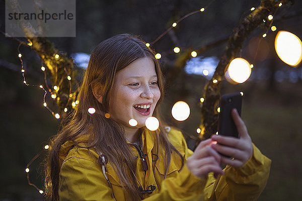 Fröhliches Mädchen benutzt Smartphone inmitten beleuchteter Lichterkettendekoration bei Nacht