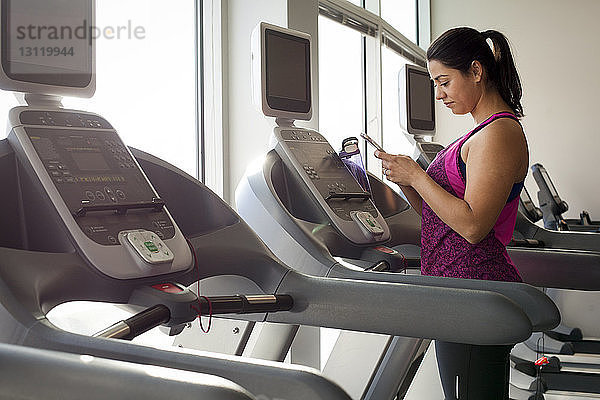 Seitenansicht einer Frau  die ein Smartphone benutzt  während sie im Fitnessstudio auf dem Laufband steht