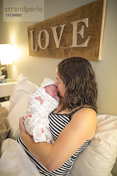 Mutter küsst neugeborene Tochter  während sie zu Hause im Bett sitzt