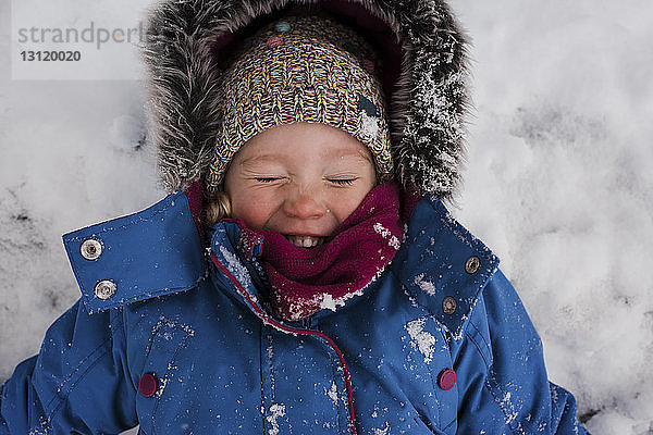 Hochwinkelaufnahme eines lächelnden Mädchens mit geschlossenen Augen  das auf einem schneebedeckten Feld liegt