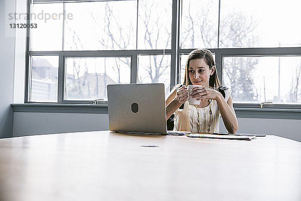 Geschäftsfrau hält Getränk in der Hand und schaut auf Laptop-Computer  während sie im Sitzungssaal sitzt