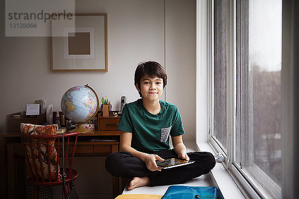 Porträt eines Jungen  der eine Tafel hält und zu Hause am Fenster auf dem Tisch sitzt