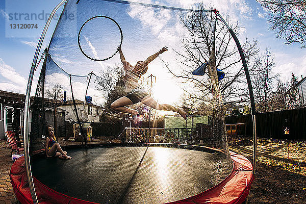 Mädchen mit Plastikreifen springt  während die Schwester auf dem Trampolin im Garten sitzt