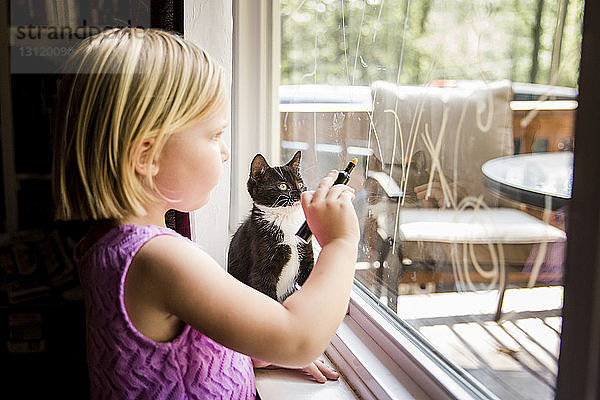 Seitenansicht eines Mädchens  das auf Fensterglas schreibt  während es zu Hause beim Kätzchen steht