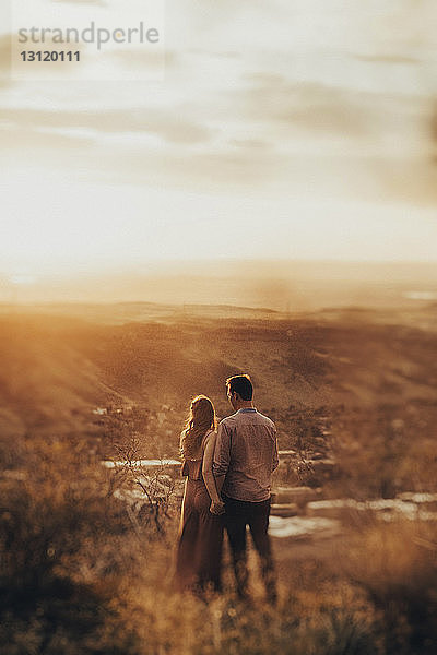 Rückansicht eines jungen Paares  das bei Sonnenuntergang auf dem Feld steht