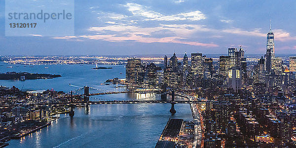 Hochwinkelansicht der Brücken von Brooklyn und Manhattan über den East River bei beleuchteter Stadtlandschaft in der Abenddämmerung