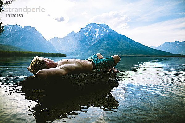 Mann ohne Hemd liegt auf Felsen im See und entspannt sich an Bergen