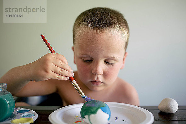 Junge ohne Hemd färbt Ostereier  während er zu Hause am Tisch sitzt