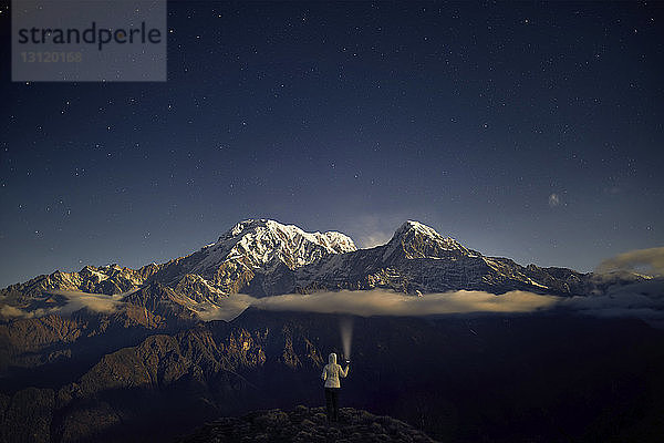 Rückansicht eines Touristen  der eine Taschenlampe hält  während er nachts auf einem Berg gegen den Himmel steht