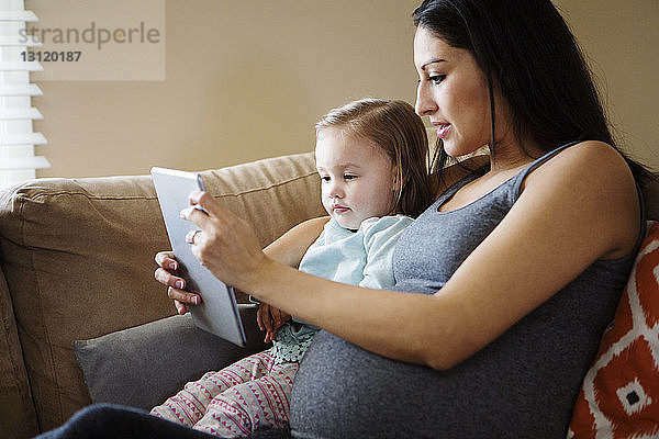 Nahaufnahme einer schwangeren Frau  die der Tochter die Tablette zeigt  während sie zu Hause auf dem Sofa sitzt