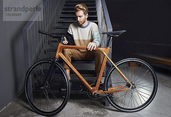 Junger Mann in voller Länge hält Fahrrad  während er zu Hause auf einer Treppe sitzt