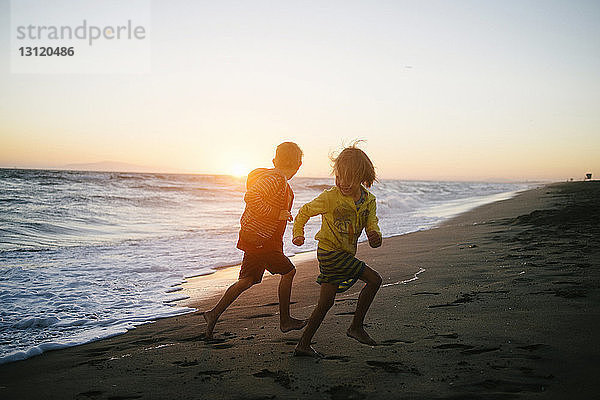 Geschwister laufen bei Sonnenuntergang am Strand gegen den Himmel