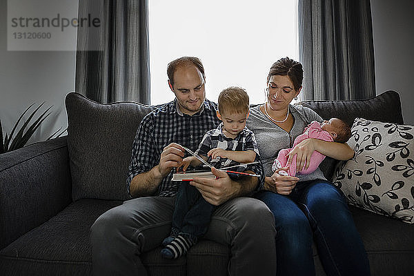 Eltern mit Kindern schauen Fotoalbum an  während sie zu Hause auf dem Sofa sitzen