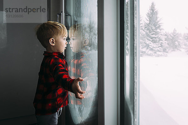 Seitenansicht eines Jungen  der durch ein Fenster schaut  während er im Winter zu Hause steht