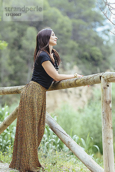 Nachdenkliche Frau in voller Länge schaut weg  während sie am Holzzaun steht
