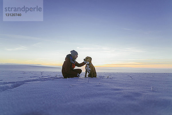 Wanderer streichelt Hund  während er bei Sonnenuntergang auf verschneitem Feld gegen den Himmel sitzt