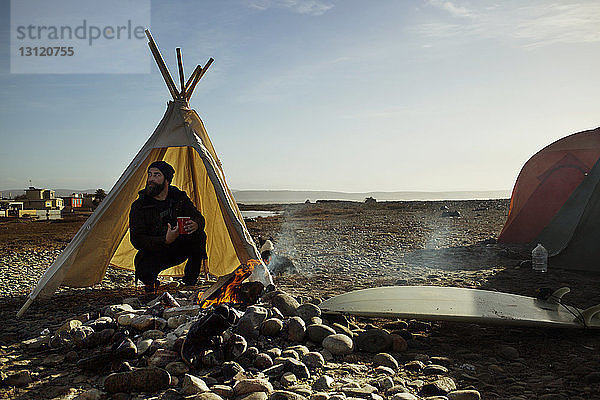 Mann mit Kaffeetasse kauert im Zelt am Lagerfeuer am Strand