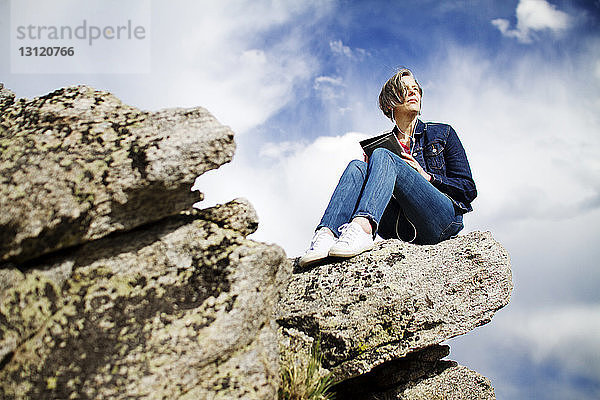 Niedrigwinkelansicht einer Frau  die Musik hört  während sie auf einem Felsen vor bewölktem Himmel sitzt