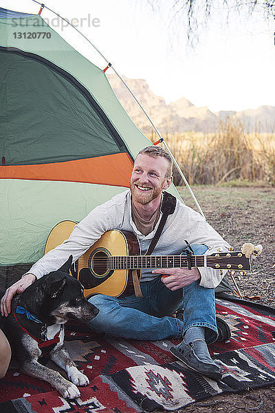 Glücklicher Mann streichelt Hund  während er Gitarre vor dem Zelt hält