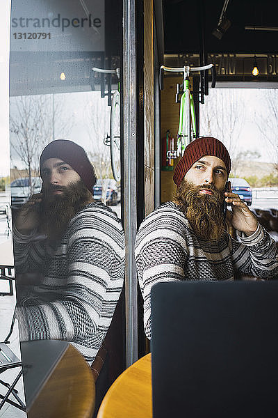Hipster-Mann antwortet auf Smartphone  während er mit Laptop-Computer auf dem Tisch im Cafe durchs Fenster schaut
