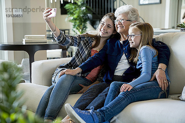 Glückliche Großmutter und Enkelinnen  die sich auf dem Sofa sitzend per Smartphone selbstständig machen