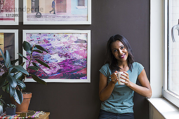 Porträt einer Frau  die eine Kaffeetasse in der Hand hält  während sie zu Hause vor einem Bilderrahmen an der Wand steht