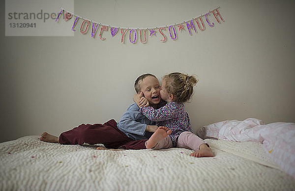 Glückliche Schwester küsst zu Hause den Bruder  während sie auf dem Bett sitzt