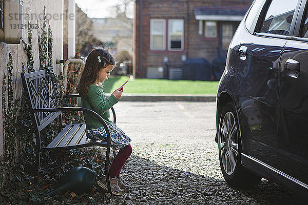 Seitenansicht eines Mädchens  das ein Mobiltelefon benutzt  während es mit dem Auto auf einer Bank am Bürgersteig sitzt