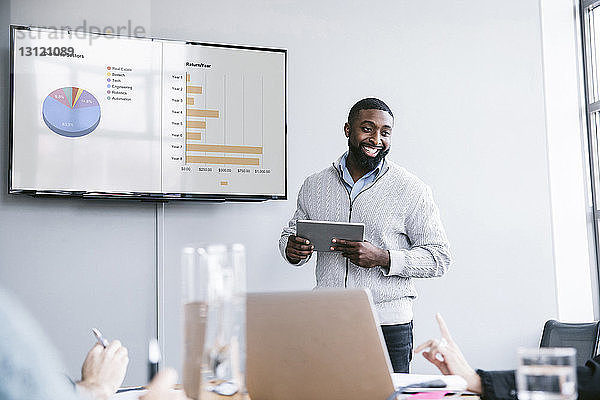 Lächelnder Geschäftsmann hält Tablet-Computer in der Hand  der bei einer Besprechung im Sitzungssaal Grafiken erklärt