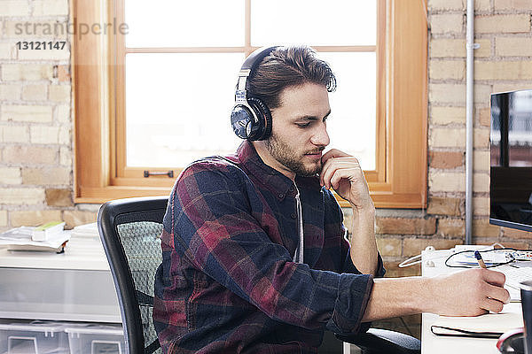 Geschäftsmann mit Kopfhörern schreibt während der Arbeit im Büro Tagebuch