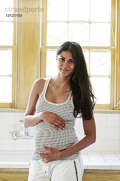 Lächelnde schwangere Frau steht zu Hause am Fenster