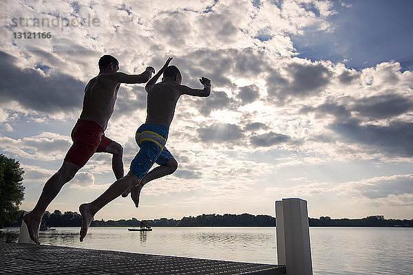 Vater und Sohn springen von der Mole am See gegen den Himmel
