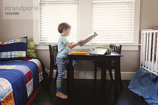 Junge hält Buch  während er im Schlafzimmer am Tisch sitzt