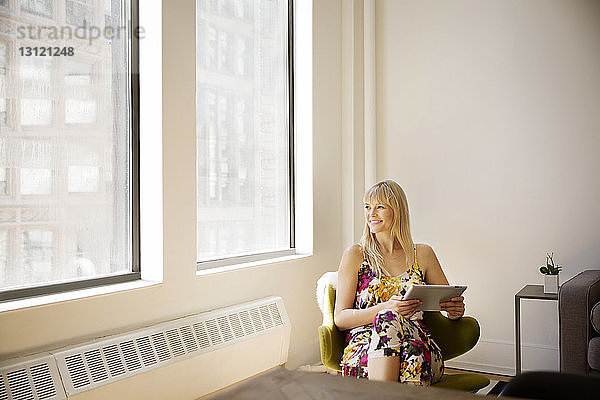 Lächelnde Geschäftsfrau hält Tablet-Computer in der Hand  während sie im Büro durch ein Fenster schaut