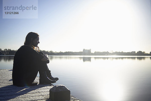 Seitenansicht einer nachdenklichen Frau  die bei Sonnenuntergang auf dem Pier über dem See vor klarem Himmel sitzt