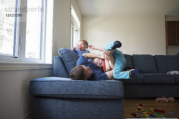 Glücklicher Vater spielt mit dem Sohn  während die Tochter zu Hause auf dem Sofa Wasser trinkt