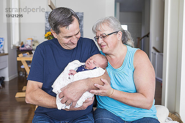 Großeltern mit süßer neugeborener Enkelin zu Hause im Wohnzimmer sitzend