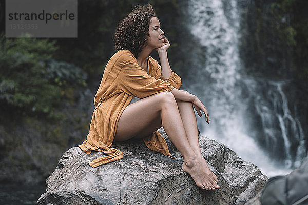 Nachdenkliche Frau schaut weg  während sie auf einem Felsen gegen einen Wasserfall im Wald sitzt